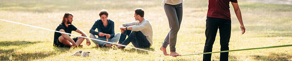 A young woman balances on a slackline in the park, a young man helps her